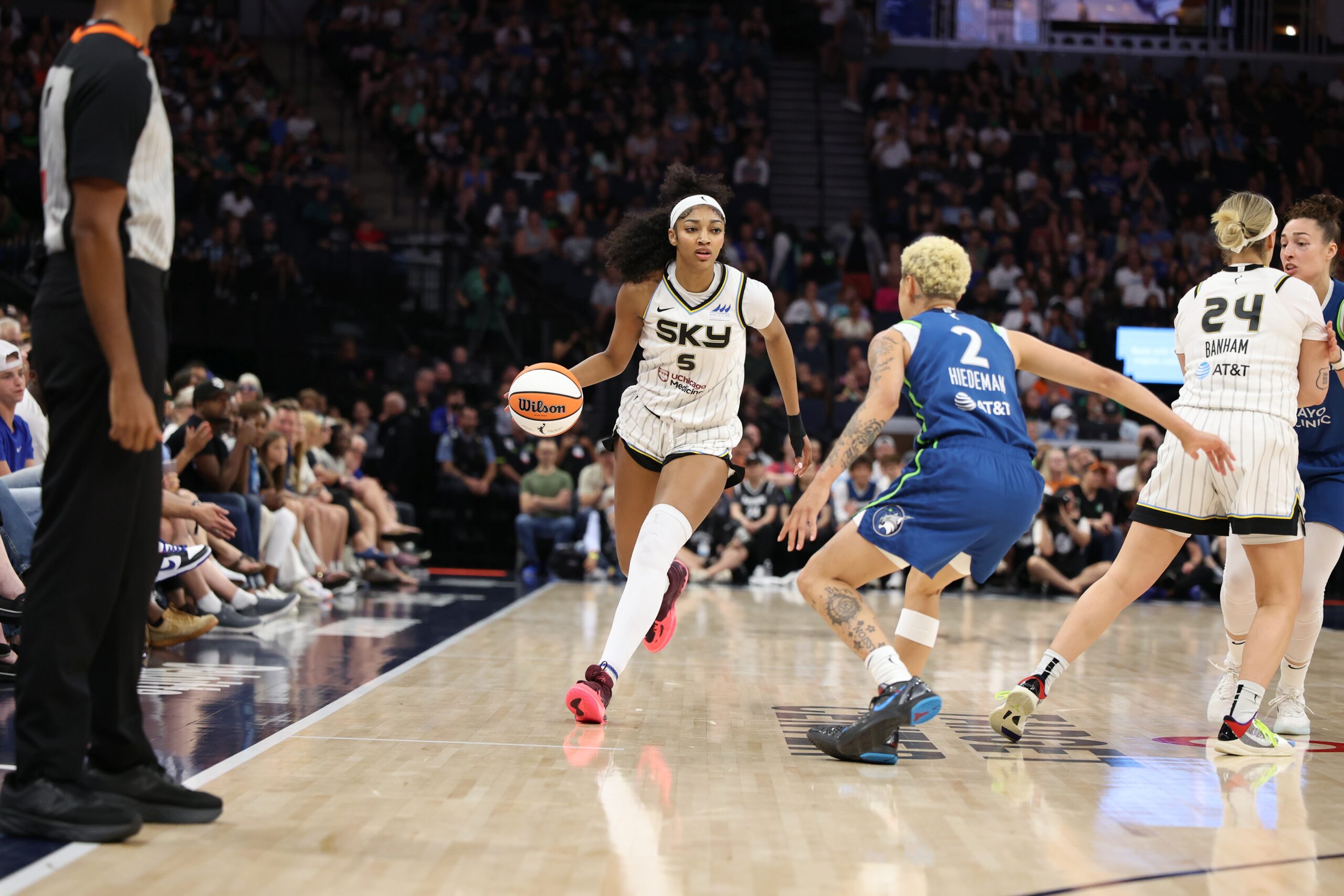 Chicago forward Angel Reese dribbles toward the viewer while Minnesota guard Natisha Hiedeman faces her in a defensive stance while shuffling backward. Next to them, Sky guard Rachel Banham is screening Lynx big Jessica Shepard away from Reese. Dozens of rows of a tightly packed crowd sit in the background and to the side of the court.