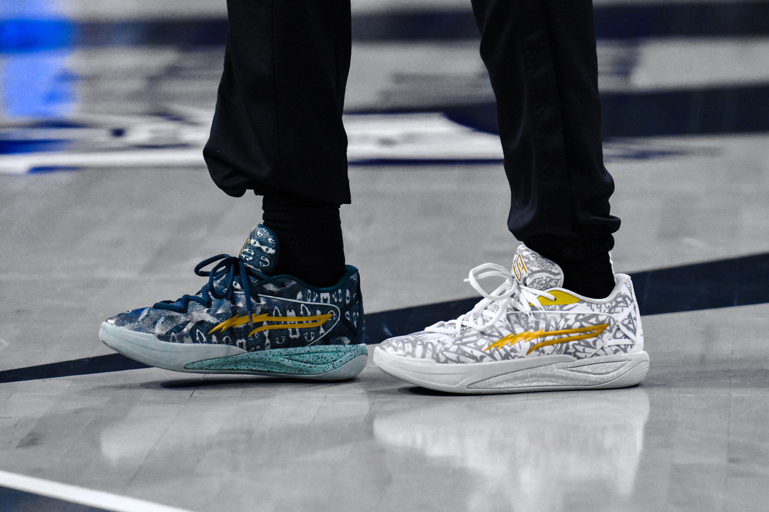 A shot of New York Liberty forward Breanna Stewart from the shins down standing on a basketball court, her black sweatpants tucked into black tube socks, wearing two shoes of different color schemes: on her left foot, white and gray designs resembling owl wings all over the sneaker, with the logo accents in gold; on the right foot, designs meant to look like the fangs and/or the outline of a snake mouth in white on a navy blue background across the shoe with the logos in gold