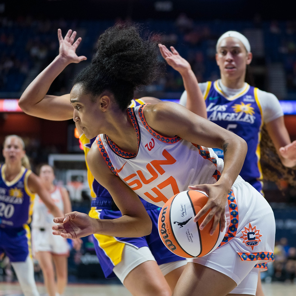 Connecticut Sun guard Leïla Lacan dribbles the ball past a Los Angeles Sparks defender who has her hands up in an attempt not to foul. Other Sparks look on from the background, in front of a blurry arena crowd.