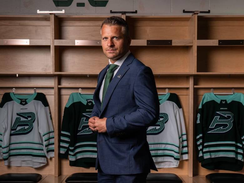 A full-body shot of Sparre in front of Fleet jerseys hanging in lockers. He is wearing a navy blue suit.