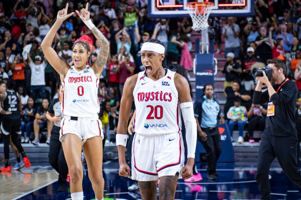 Washington Mystics guard Brittney Sykes flexes downward and yells in celebration. Center/forward Shakira Austin stands behind her, holding her hands above her head and making a "W" with her fingers.