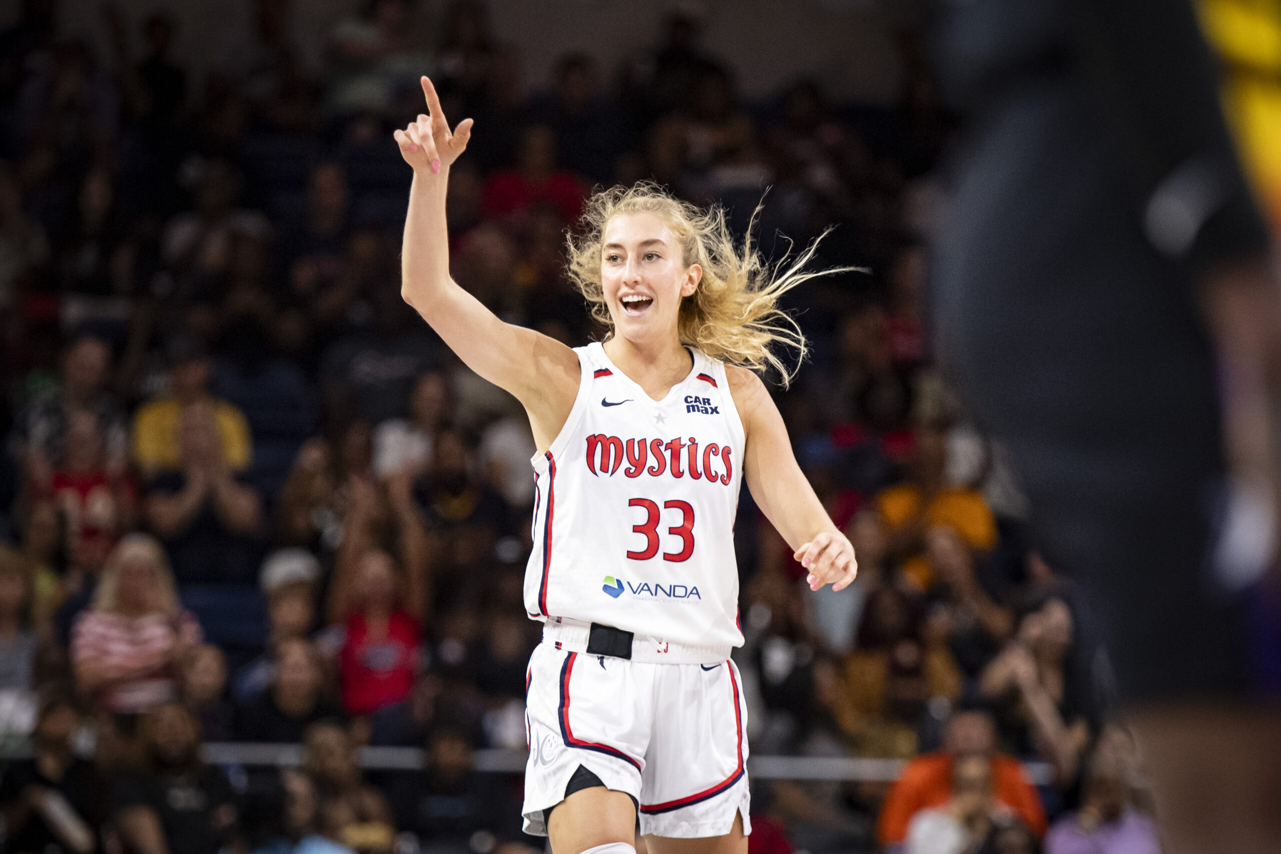 Washington Mystics guard Lucy Olsen raises her right hand and points up ahead of her. She is alone in the frame, and her mouth is slightly open, like she might be saying something.