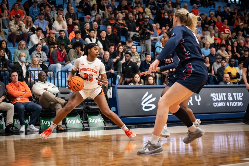 Princeton guard Madison St. Rose handles the ball with her left hand inside the 3-point line. Her legs are spread wide as she tries to shed two Penn defenders, and she is looking up and to her left.
