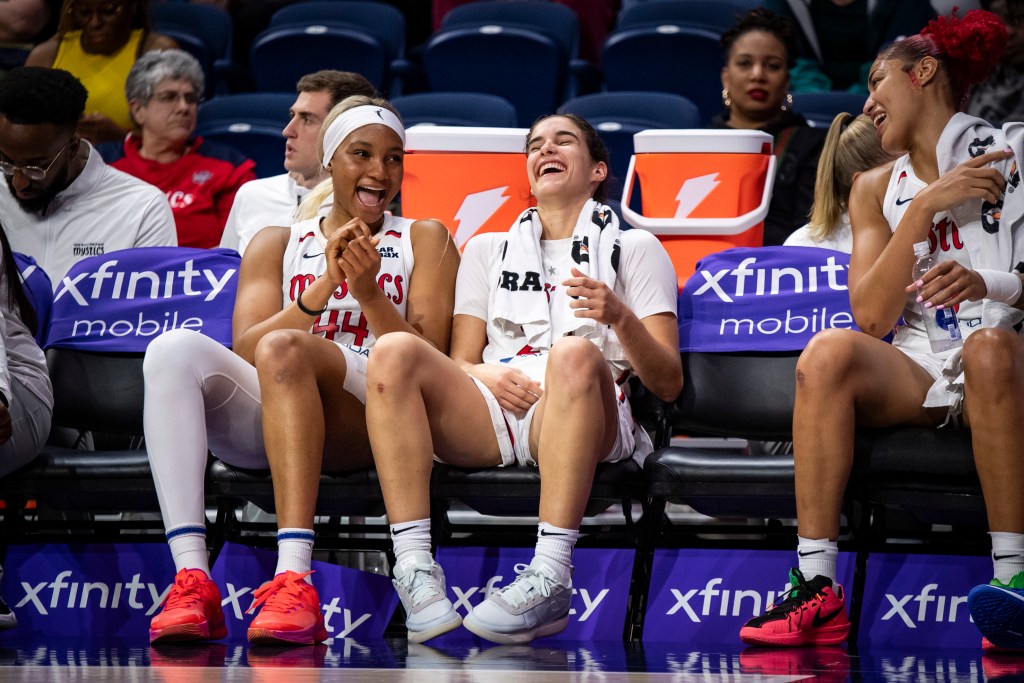 Washington Mystics forward Kiki Iriafen, guard Sonia Citron and center/forward Shakira Austin sit on the bench during a game. Iriafen is smiling with her mouth open and her hands together in front of her. Citron is laughing with her head tilted slightly up. Austin is looking toward them and holding a water bottle in her left hand.