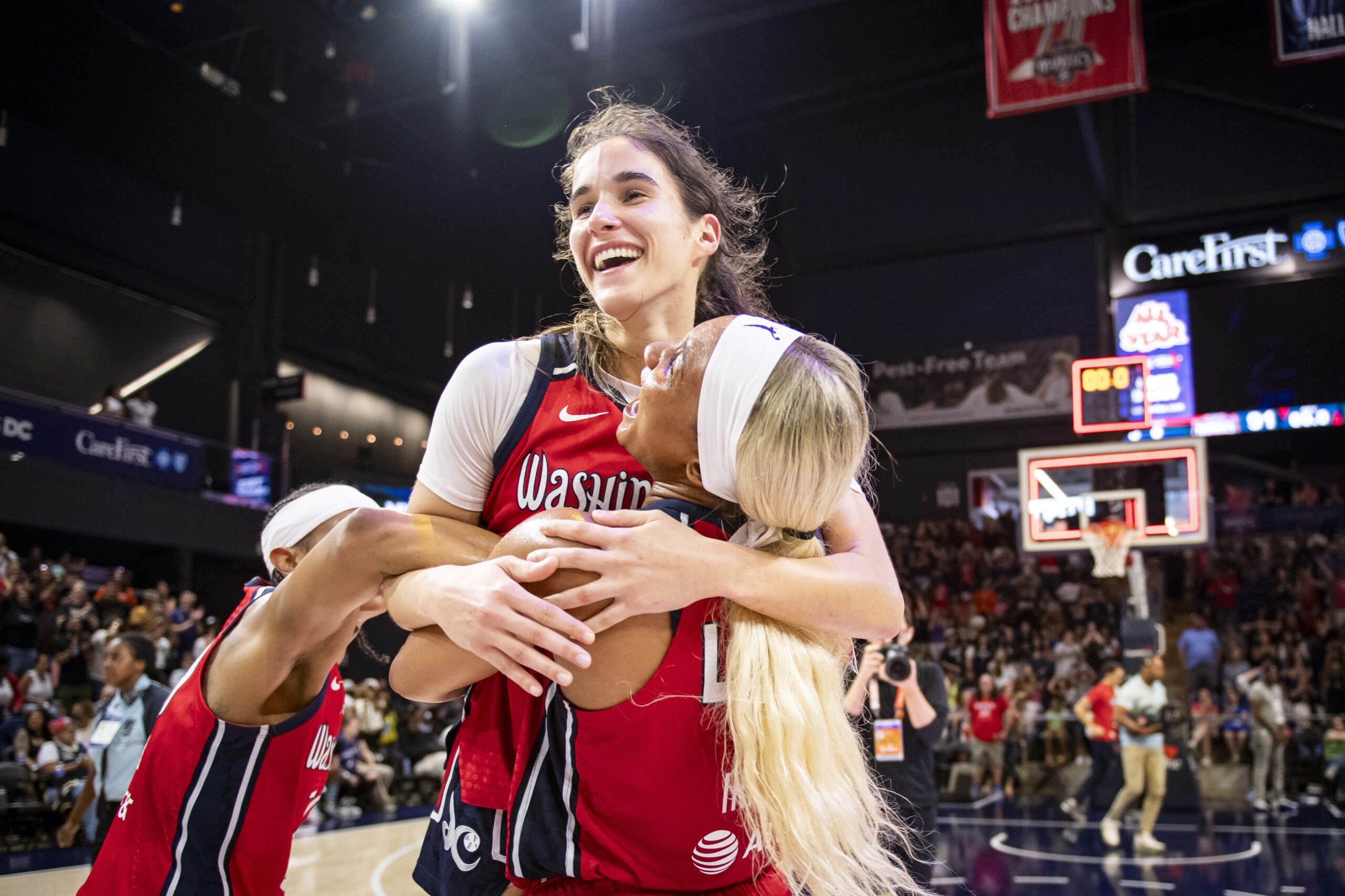 Washington Mystics forward Kiki Iriafen shouts as she picks up guard Sonia Citron. Citron is beaming and has both arms around Iriafen's shoulders. Guard Brittney Sykes also has one arm tangled around them.