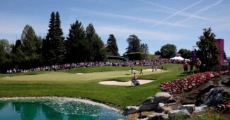 A view of the green of the 5th hole at the Evian Championship in France. A bright blue pond of water is in the lower left corner of the shot. Pink flower beds and rocks line the right side of the picture. Tall trees stretch behind the green, and the grandstand of fans sits in the back right.