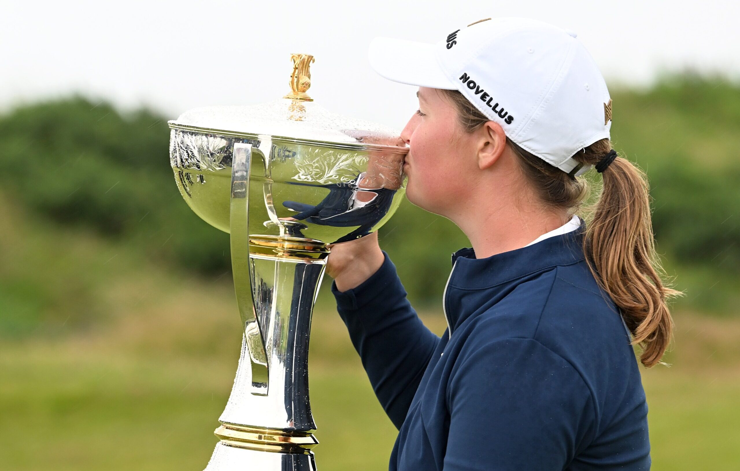 Lottie Woad, winner of the 2025 ISPS HANDA Women's Scottish Open, holds up the large shiny trophy to her right and kisses the bowl. The green gorse and bushes of Dundonald Links are in the backdrop.