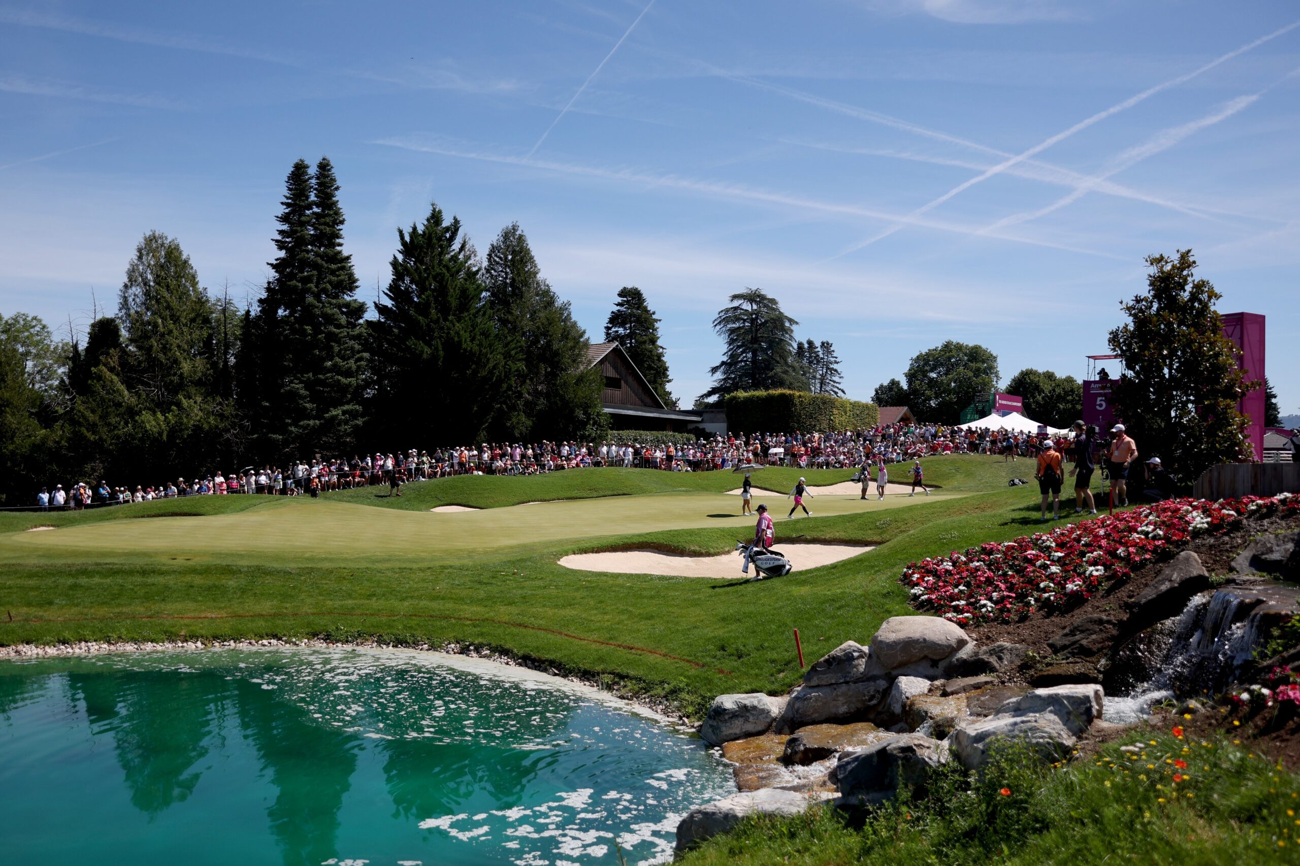 A view of the green of the 5th hole at the Evian Championship in France. A bright blue pond of water is in the lower left corner of the shot. Pink flower beds and rocks line the right side of the picture. Tall trees stretch behind the green, and the grandstand of fans sits in the back right.
