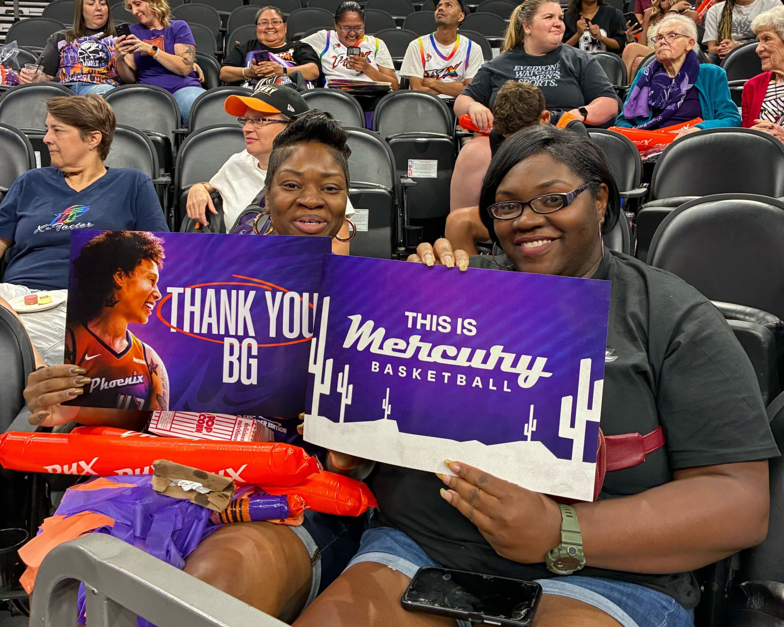 Two Phoenix Mercury fans holding signs that say "Thank You BG" (left) and "This Is Mercury Basketball" (right) with purple and orange graphics and photos of Brittney Griner.