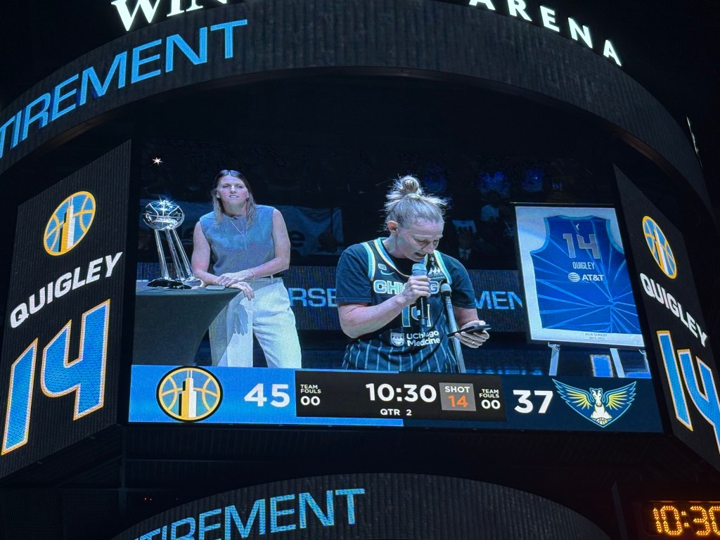 A blonde woman with her hair in a bun (Vandersloot) is in the foreground speaking into a microphone. Behind her to the left, another woman (Quigley) stands next to a trophy. On the other side of the blonde woman is a framed jersey with the number 14 on it.