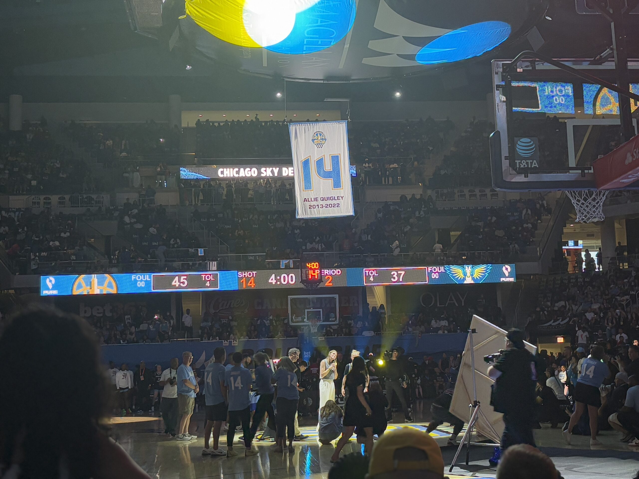 A large white banner with the number 14 on it hangs from the ceiling of a large basketball arena. Right under it, in a beam of light, stands a woman surrounded by a crowd of people in shadows.