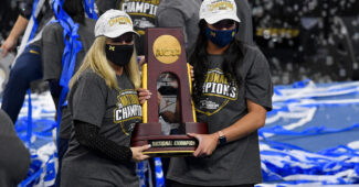 Two women, including Michigan head coach Bev Plocki, in masks accept a trophy.