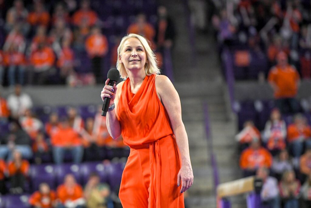 Wearing an orange dress, Clemson head coach Amy Smith holds a microphone before a large arena crowd.