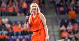 Wearing an orange dress, Clemson head coach Amy Smith holds a microphone before a large arena crowd.