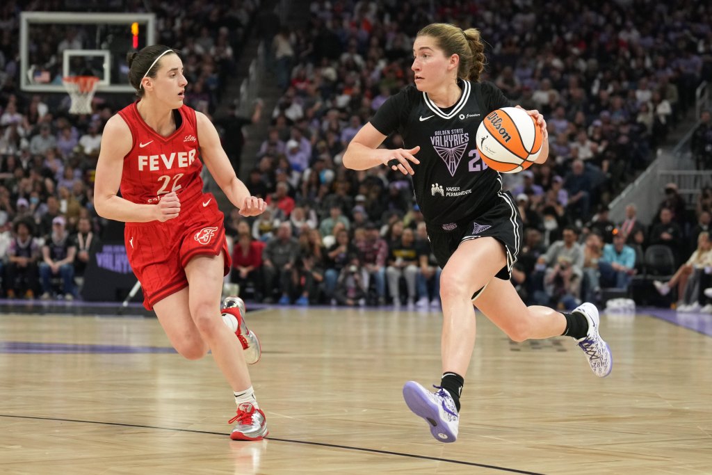 Golden State Valkyries guard Kate Martin dribbles against Indiana Fever guard Caitlin Clark. They are shown from the side, and a packed but out-of-focus crowd is in the background behind them.