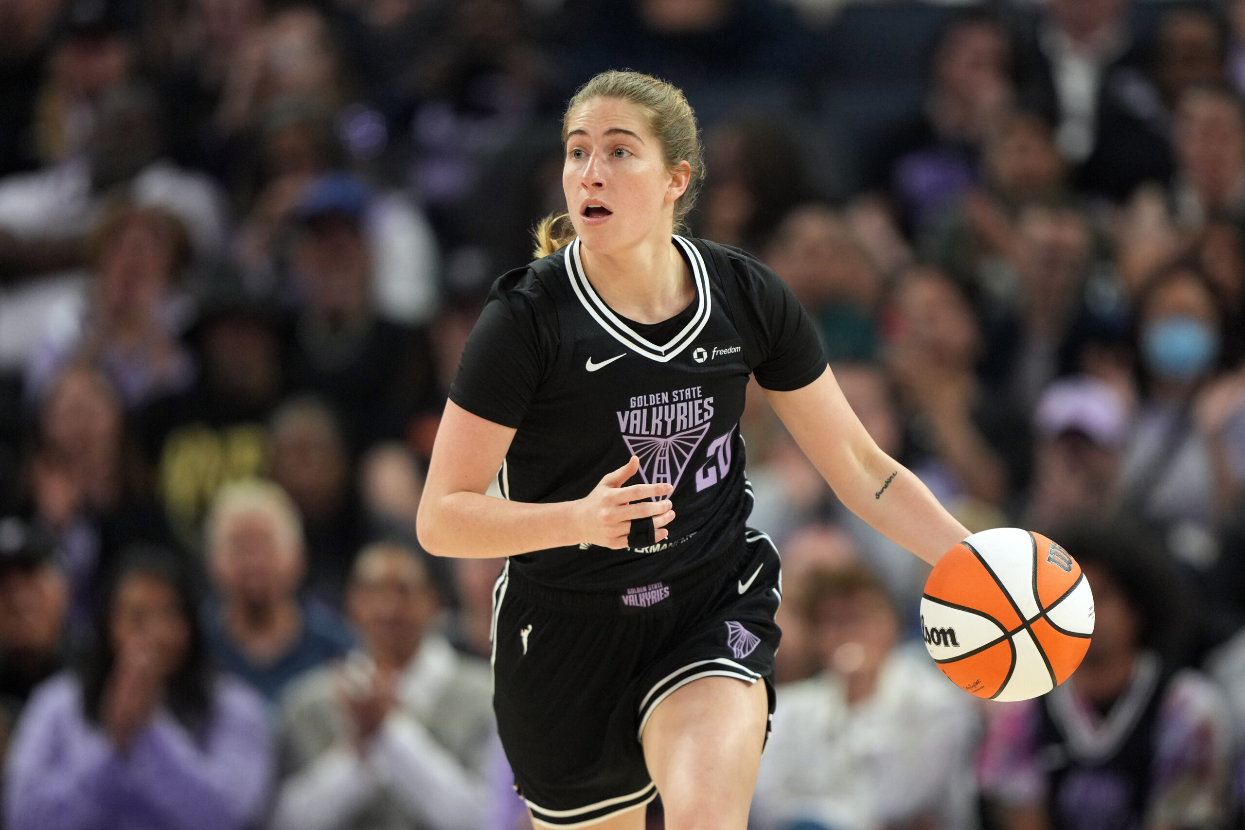 Golden State Valkyries guard Kate Martin dribbles the ball. She is shown from the front, and a packed but out-of-focus crowd is in the background behind her.