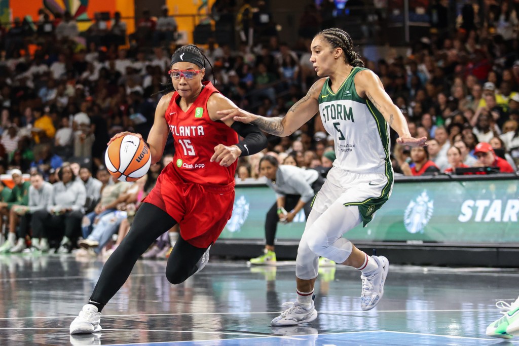 Two players battle for the ball during the game. The player on the left dribbles the ball and wears a red Atlanta Dream jersey. The player on the right attempts to steal the ball and wears a green and white Seattle Storm jersey. 