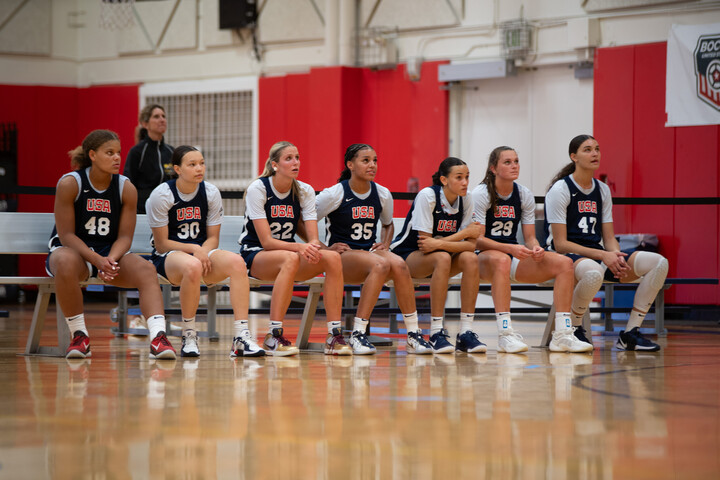 Seven members of the United States Under-19 team sit on the bench during practice and watch the action on the court.