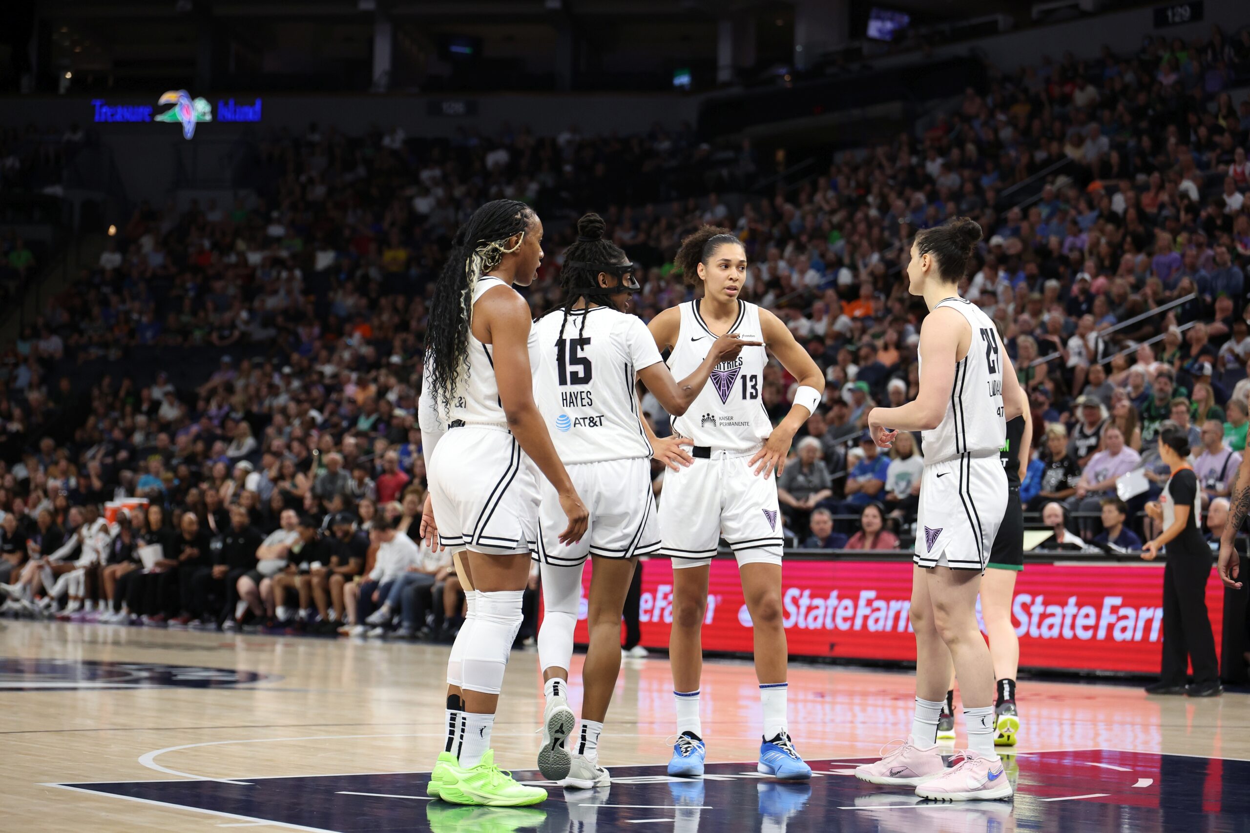 Golden State Valkyries players Kayla Thornton, Tiffany Hayes, Janelle Salaün, and Cecilia Zandalasini huddle up in the paint during a break in play. A tightly packed crowd fills the seats of an arena in the background.