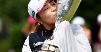 Ayaka Furue holds the Amundi Evian Championship trophy up to her left and kisses the side of it. Her hands are wrapped in the Japanese flag draped over her shoulders.