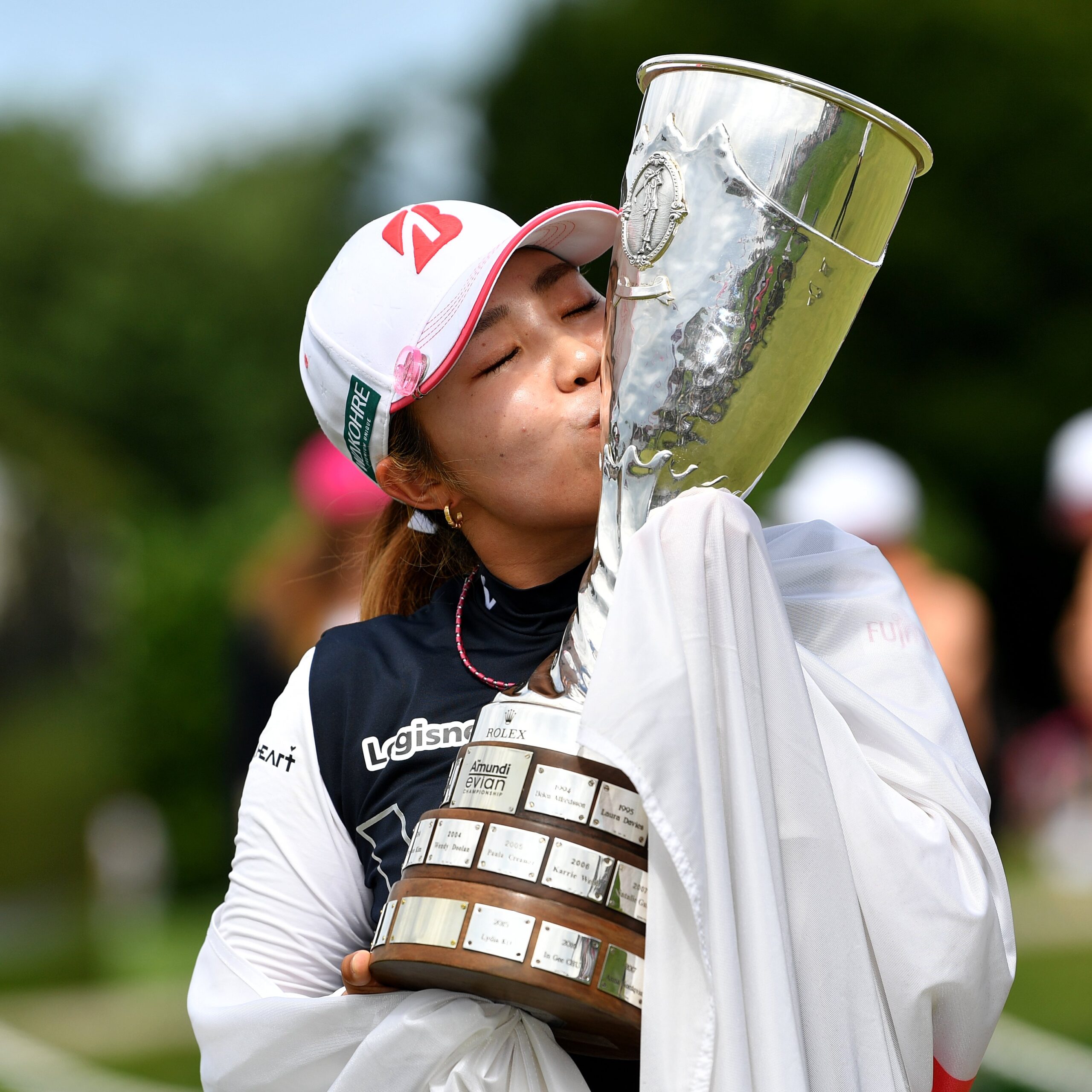 Ayaka Furue holds the Amundi Evian Championship trophy up to her left and kisses the side of it. Her hands are wrapped in the Japanese flag draped over her shoulders.