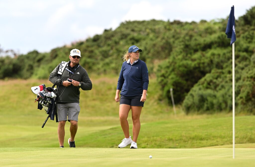 Lauren Coughlin, winner of the 2024 ISPS HANDA Women's Scottish Open, approaches the green. She looks to the right side of the picture. Her caddie is to the left of her and carries her golf bag over his right shoulder. The flag on the green is in the front right of the frame.