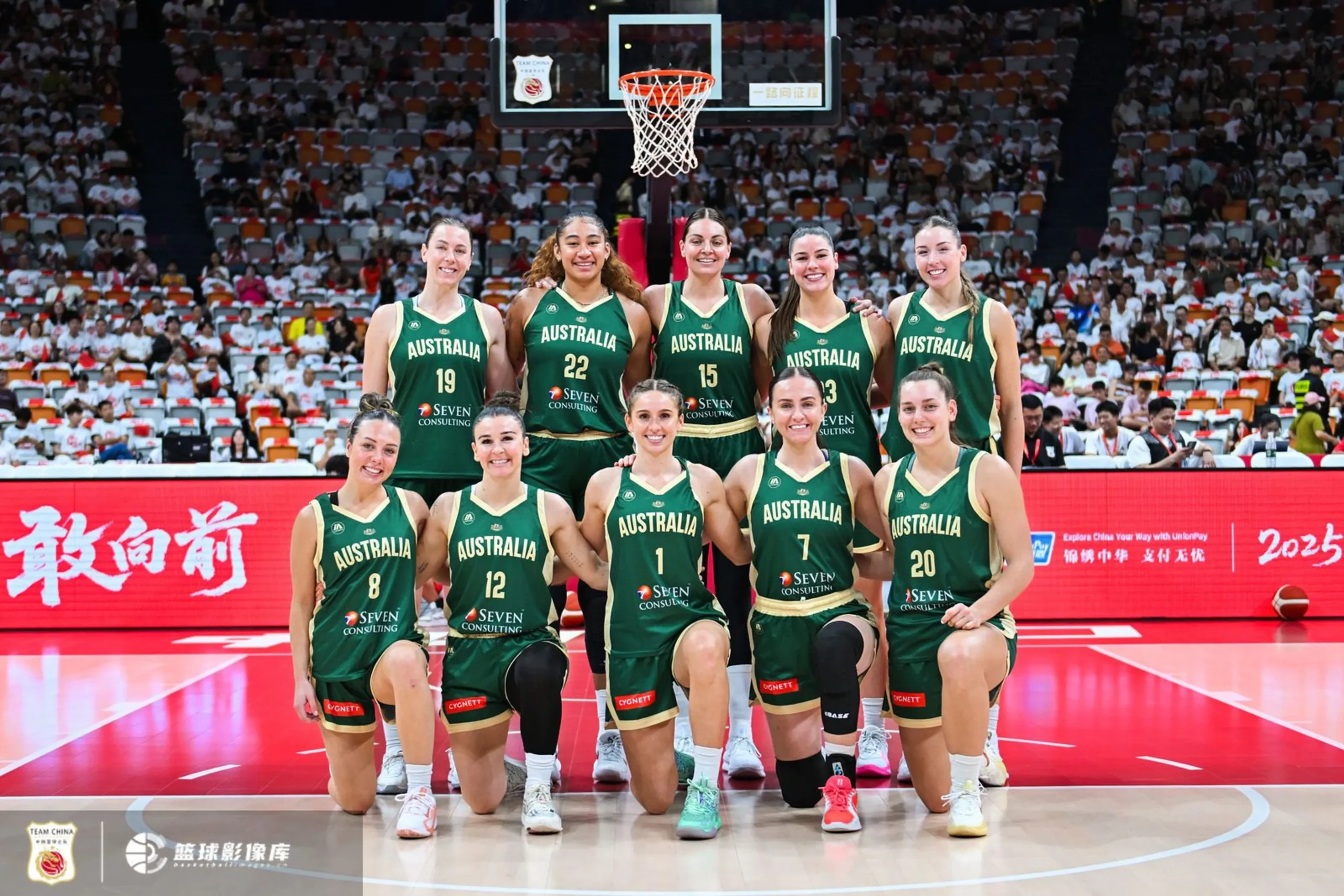 Two rows of Australian basketball players take a team photo before a game. They are on the free throw line with the rim and crowd in the background.