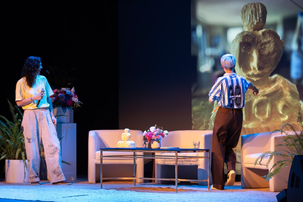 Megan Rapinoe and Sue Bird, facing backwards, on stage presenting during a live taping of their podcast. Behind them on a screen is a statue made out of butter.