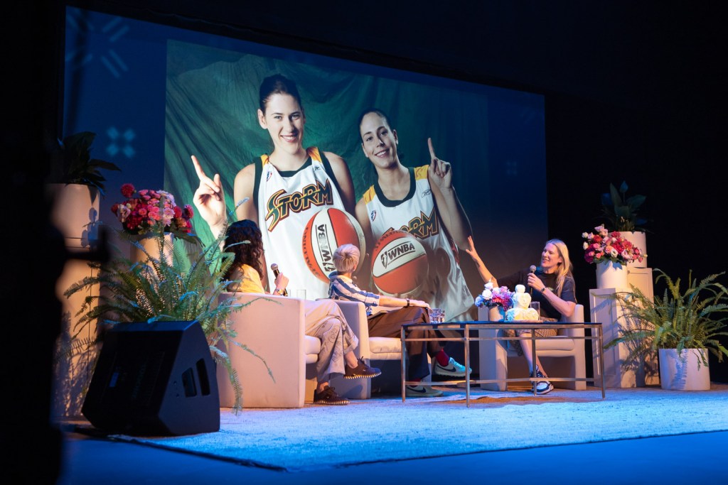 Sue Bird, Megan Rapinoe, and Lauren Jackson sit on stage during a live taping of Bird and Rapinoe's podcast. They sit on couches, holding microphones while a banner is displayed behind them that features Bird and Jackson from their playing days on the Seattle Storm.