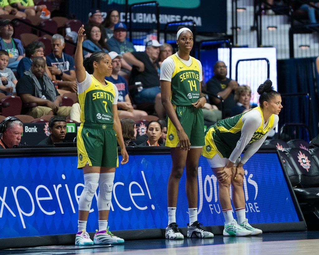 Three Seattle Storm players wait to check into a game. Center Dominique Malonga loosely tugs at her jersey, guard Zia Cooke makes the 3-point sign with her right hand, and forward Alysha Clark watches the game with her hands on her knees.
