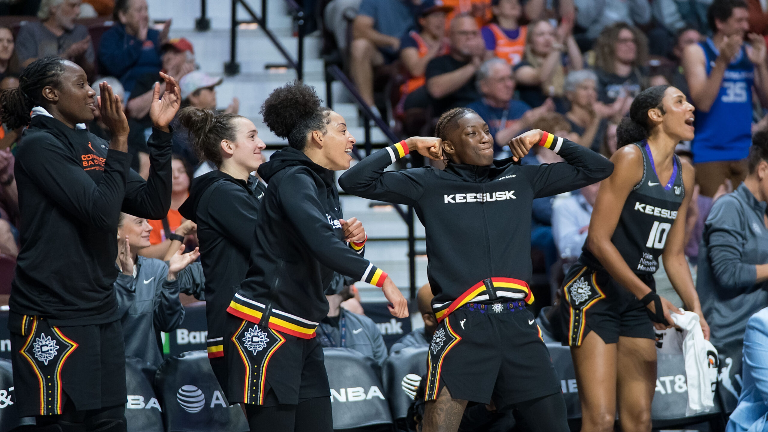 Connecticut Sun players celebrating on the bench. One player is flexing both arms and the other players are smiling and cheering for their teammates.