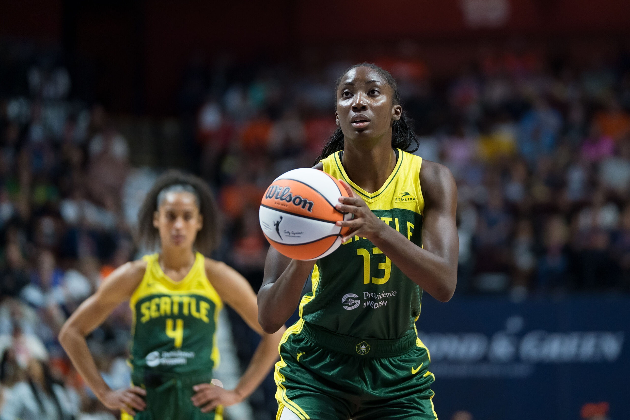 Ezi Magbegor stoically raises her arms to take a free throw. Skylar Diggins stands behind her, blurry, with her hands on her hips.