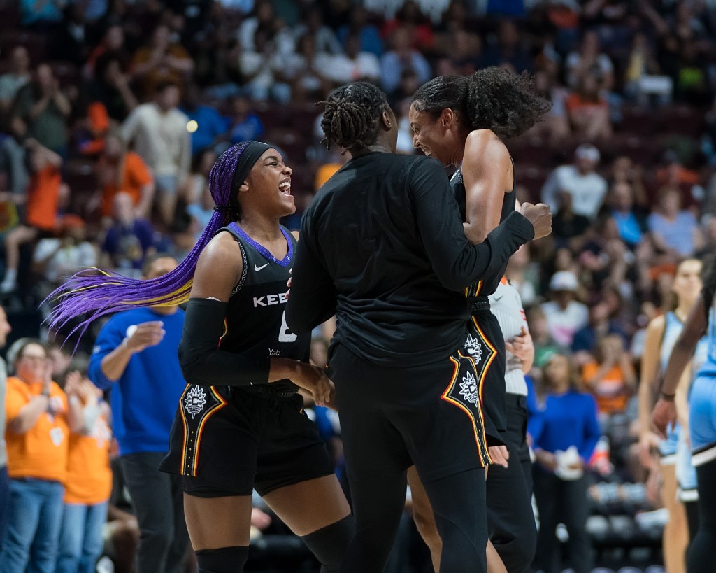 Three players laugh on the court together as they celebrate a play. They are in black uniforms with orange piping on the sides.