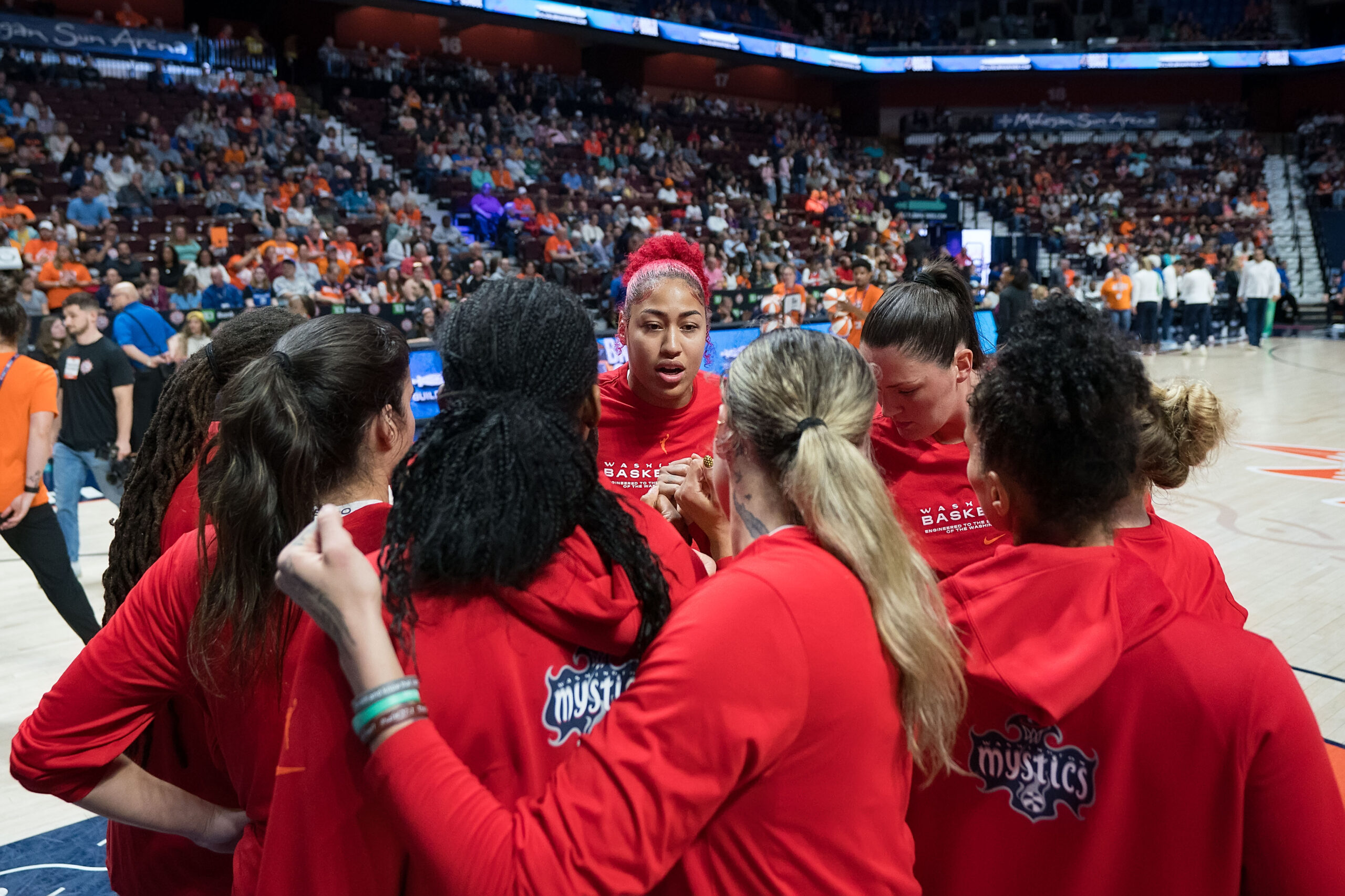 Washington Mystics players huddle in the paint before a game. They are wearing bright red warmup gear.