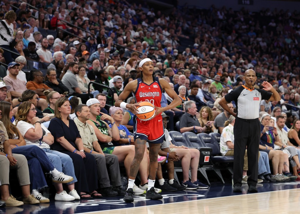 Washington Mystics guard Brittney Sykes stands on the sideline just in front of a row of fans, holding the ball in both hands. She scans the court, looking for a player to pass to, as an official stands next to her counting the seconds.
