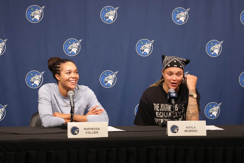 Napheesa Collier dressed in light blue sits next to Kayla McBride dressed in black, they sit in front of a dark blue backdrop and have big smiles on their faces