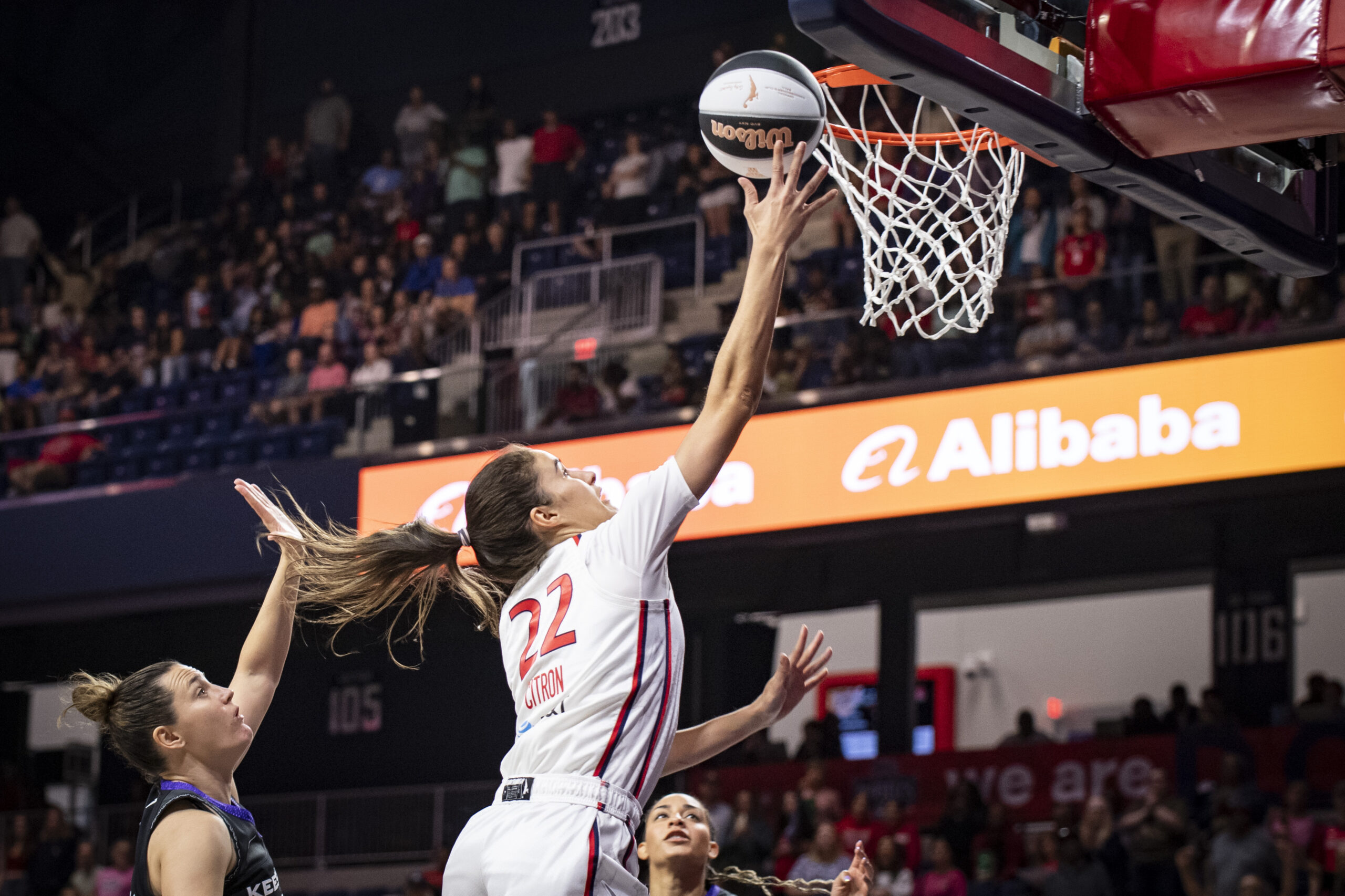 Washington Mystics guard Sonia Citron is shown in midair as she shoots a layup. Her right hand is very close to the rim, and the ball is just off her fingertips. Two Connecticut Sun defenders are visible in the frame, but neither is in position to challenge the shot.