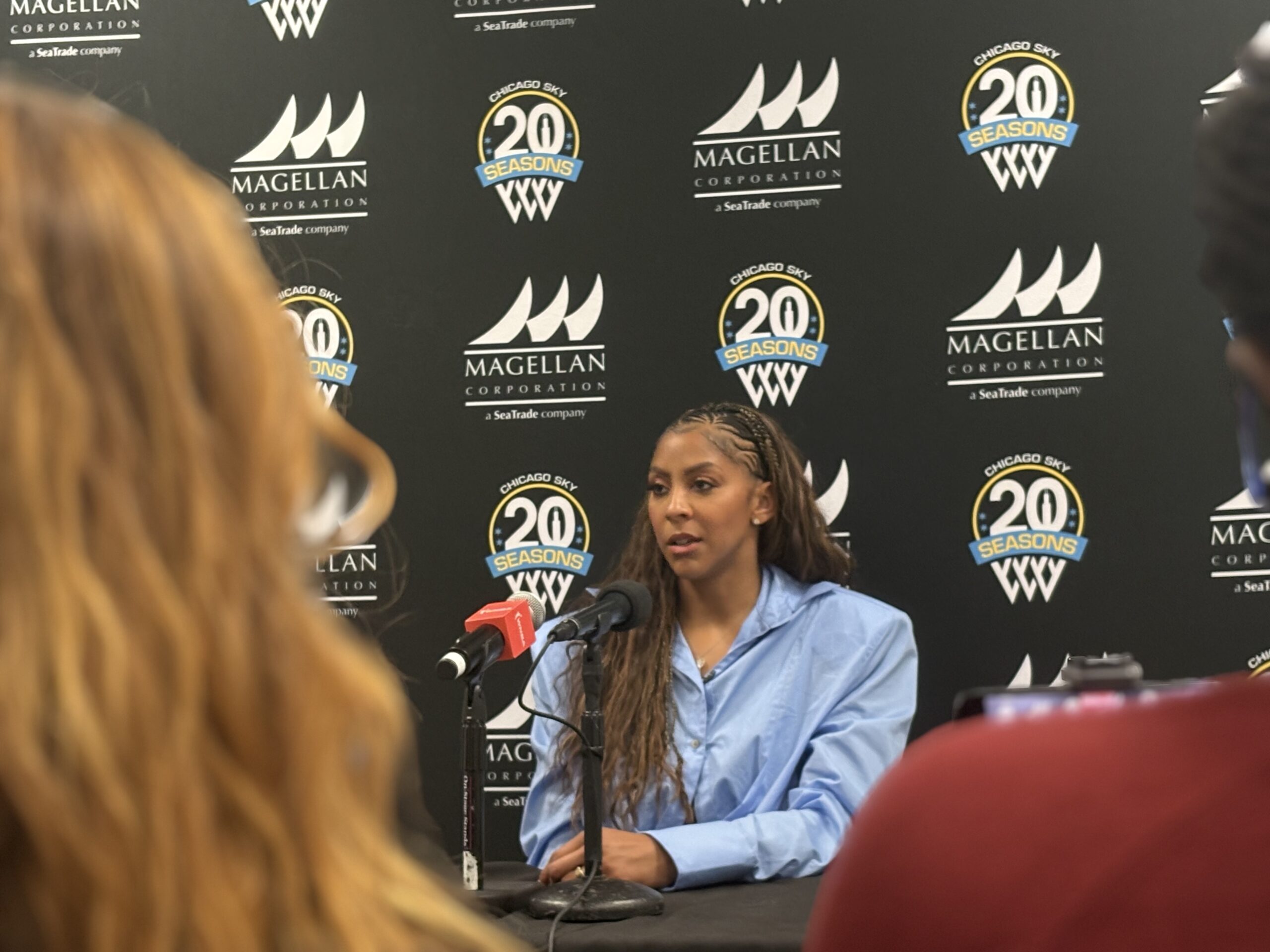 Candace Parker is sitting in front of a black, blue and white step and repeat with Chicago sky branding as she speaks into a microphone to the media.