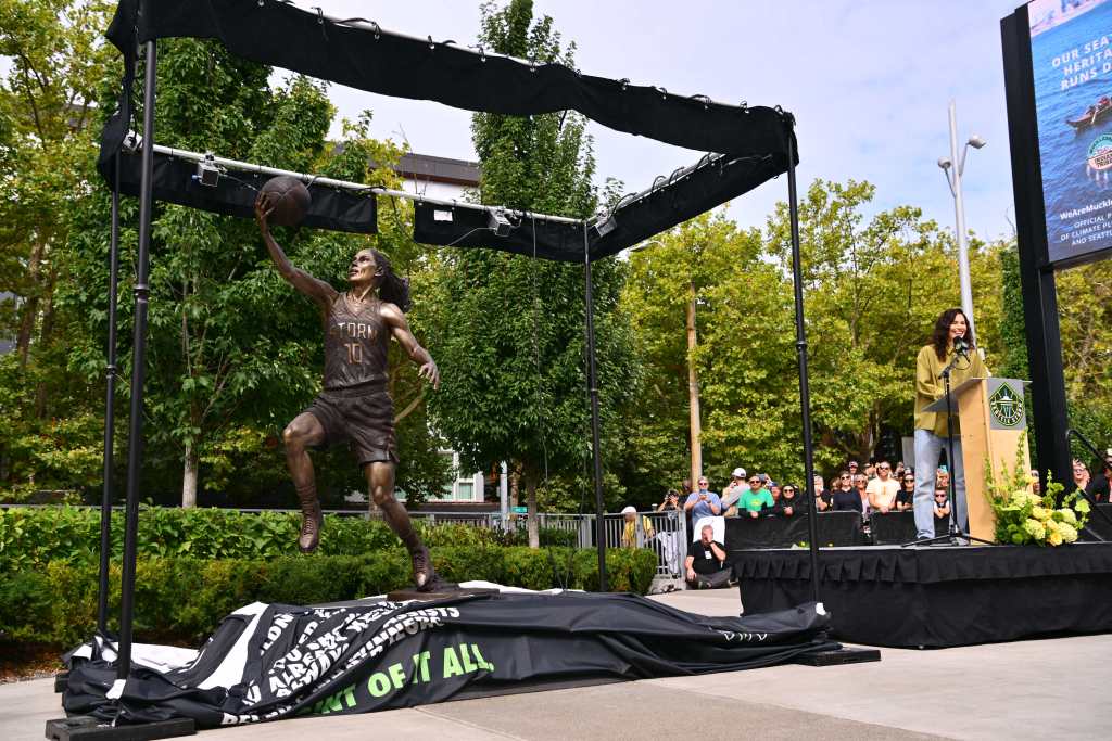 Sue Bird stands to the right at a podium in front of a statue of her making a layup that had just recently been unveiled. A crowd is gathered behind her to hear her speak.