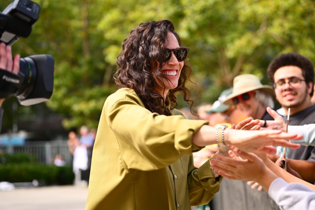 Sue Bird smiles in a green jacket in sunglasses while she stands outside at her statue unveiling, shaking hands and waving hello to fans and supporters who showed out for the event.