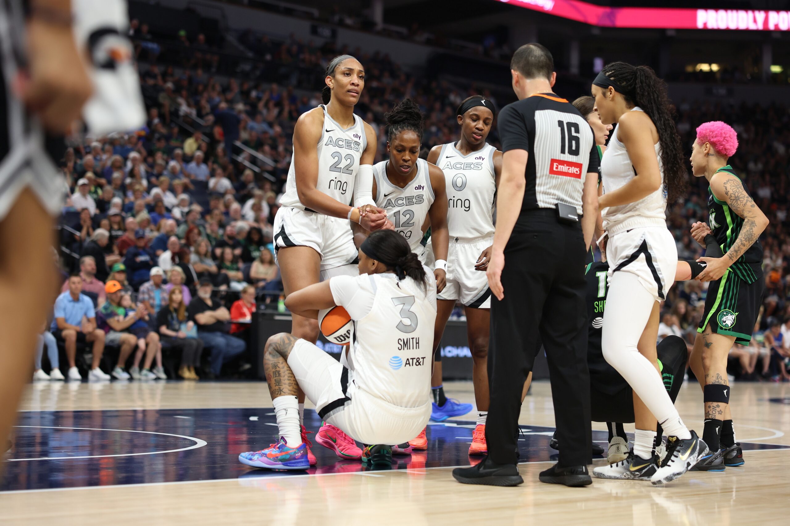 A group of Las Vegas Aces players including A'ja Wilson, Chelsea Gray, Jackie Young and Aaliyah Nye gather in the paint while Wilson helps pick NaLyssa Smith pick herself up off the ground. A referee stands next to them looking on, while a pair of Minnesota Lynx players are talking to each other behind the group. Packed arena stands are out of focus in the background.