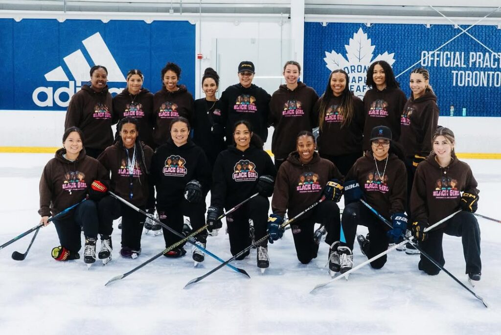 A group of women from the Black Girls Hockey Club pose in two rows on the ice for a photo. They are all smiling, and the lower row is holding hockey sticks.