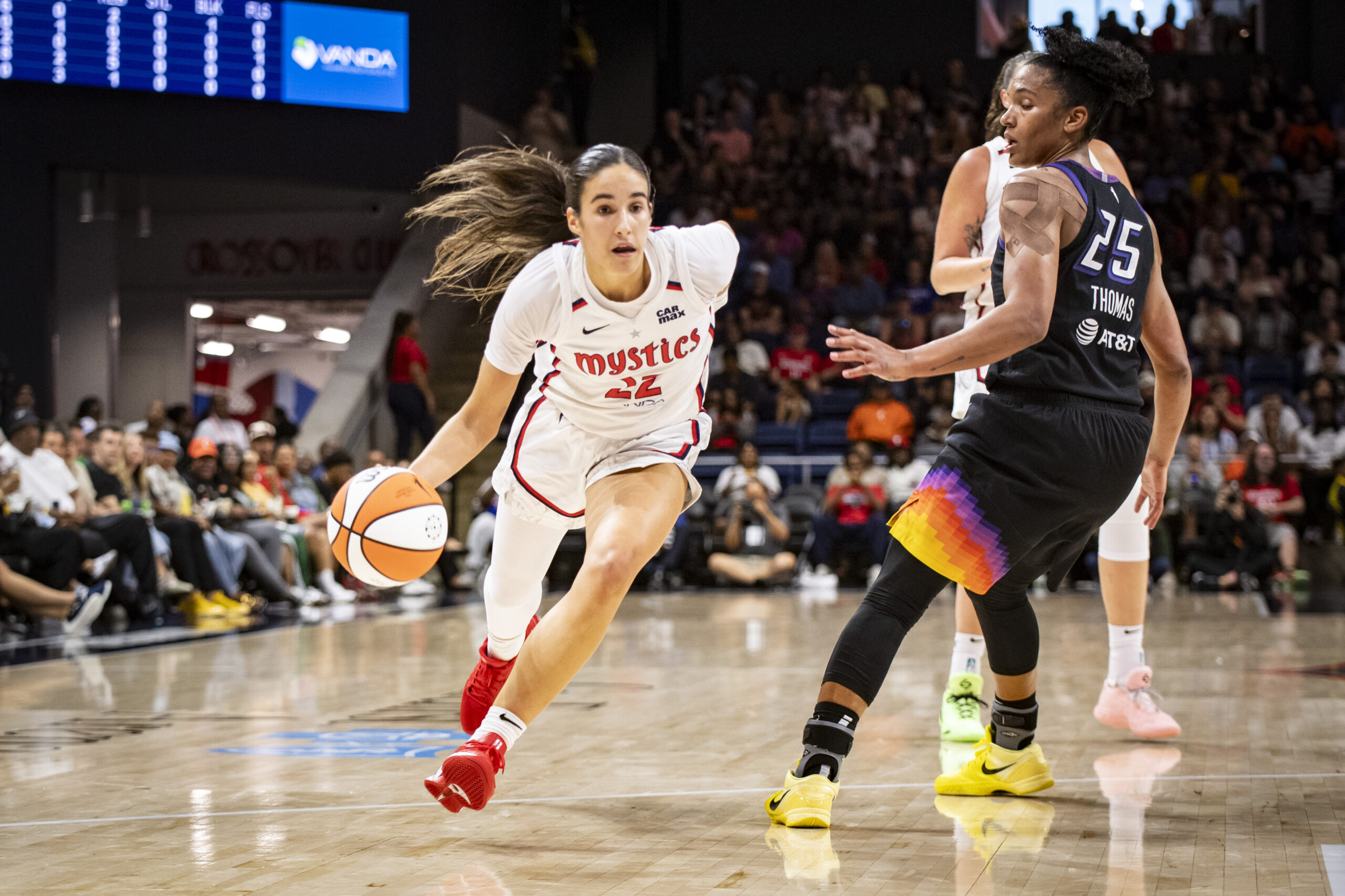 Wasington guard Sonia Citron dribbles toward the rim past Alyssa Thomas, who is trying to turn and follow. Another Mystics player stands behind Thomas, while a packed crowd on the sideline and endline watch on out of focus in the background.