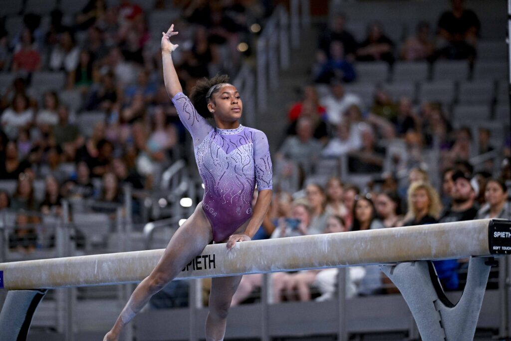 A gymnast in a purple leotard raises a hand over her head while straddling the balance beam. 