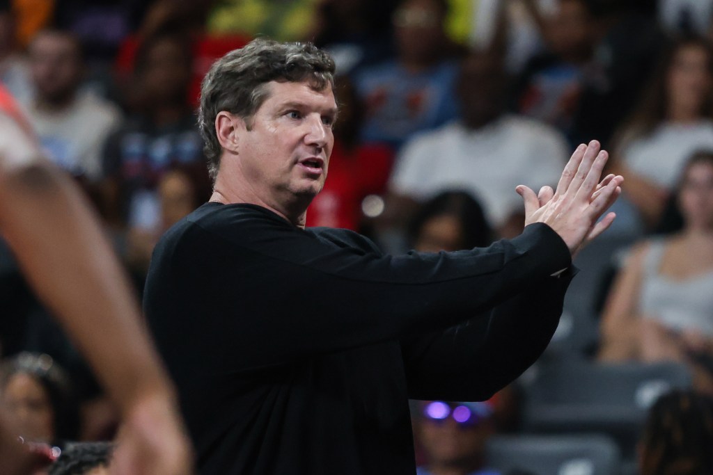 Atlanta Dream head coach Karl Smesko stands on the sidelines in a black sweater. He is coaching his team and gesturing at a play using his hands. 