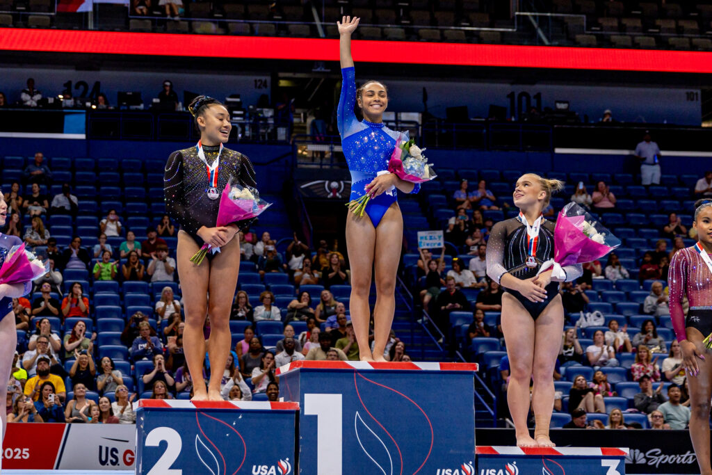 The podium at Xfinity National Gymnastics Championships, with a gymnast in a blue leotard standing in the first place position raising an arm.