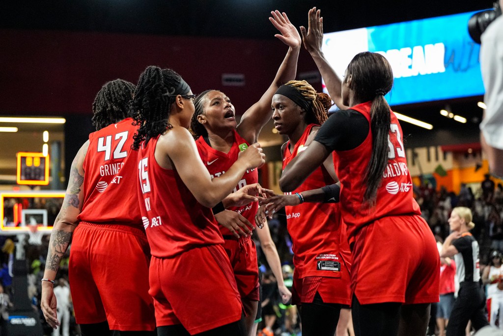 A group of Atlanta Dream players huddle on the court during a game. They are all wearing red jerseys and two players are High-fiving.