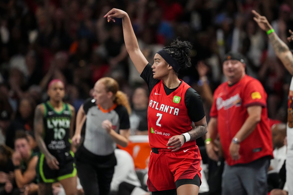A player on the Atlanta Dream gestures for the ball during a game. She is wearing a red Atlanta Dream uniform. A referee, a player on the opposing team, and fans are blurred in the background. 