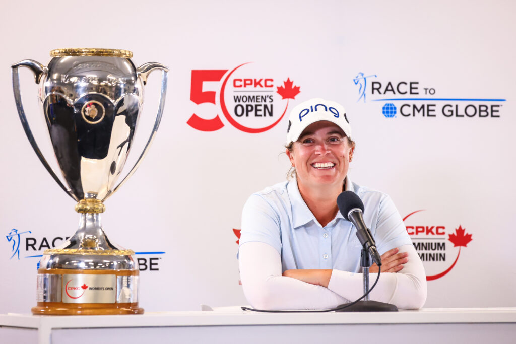2024 CPKC Women's Open champion Lauren Coughlin sits at the press conference table with her arms crossed and elbows on the table. She smiles as she talks into the mic. The CPKC Women's Championship trophy sits on the table to her right.