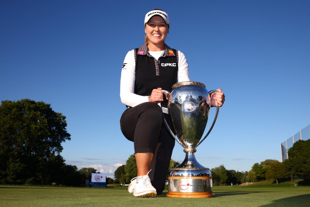 Brooke Henderson, winner of the 2025 CPKC Women's Open, kneels on the green with her right knee up and smiles at the camera. The tournament trophy sits on the ground to her left, and both of her hands grip the handles of the trophy.